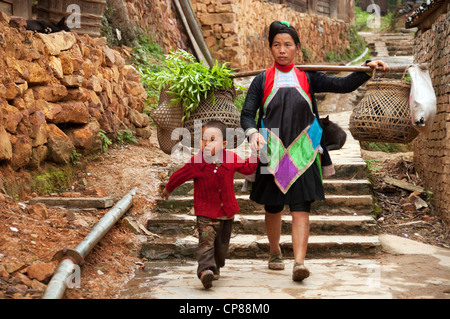 Une femme Miao basha transportant sur ses épaules avec des paniers de légumes et un enfant dans un village, Chine Banque D'Images