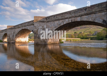 Soirée ensoleillée, vue sur la rivière du vieux pont voûté en pierre, reflétée dans les eaux peu profondes, l'eau claire de la rivière Wharfe, Tonbridge, Yorkshire, Angleterre, Royaume-Uni. Banque D'Images