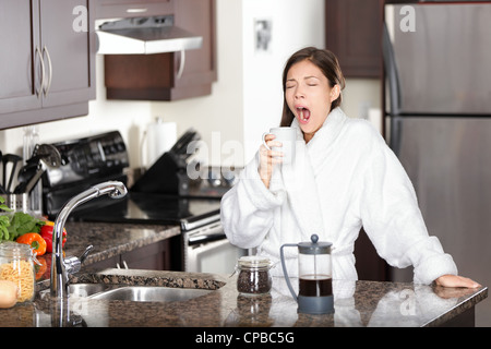 Jeune femme endormie multiraciale fatigué en peignoir de bâiller pendant que holding Coffee cup in kitchen Banque D'Images