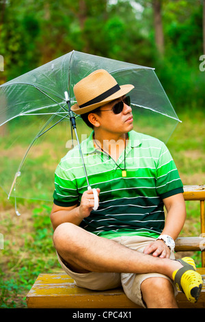 Asian young man holding an umbrella et assis sur banc de parc Banque D'Images