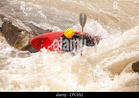 Un kayakiste dans un kayak rouge vif manœuvre à travers des rapides d'eau vive mousseux et barattant. La personne porte un casque jaune et une combinaison noire, saisissant une pagaie avec une main alors que le kayak s'incline brusquement Banque D'Images