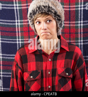 La moue sceptique jeune femme portant une chemise de bûcheron à carreaux et fur hat. Funny portrait contre un fond à motifs Banque D'Images