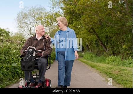 Heureusement mariée mature couple en promenade dans la campagne alors que l'un conduit un scooter de mobilité électrique motorisé Banque D'Images