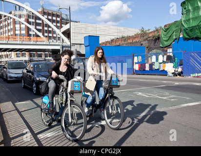 Deux jeunes femmes sur des vélos loués, attendant à un feu de circulation, Shoreditch, Londres, Angleterre, Royaume-Uni. Banque D'Images