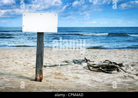 Tableau blanc vierge sur poteau en bois sur une plage de sable fin, ciel nuageux et mer en arrière-plan Banque D'Images