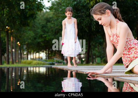 Fille assise en bord d'étang touchant la surface de l'eau, une autre fille en arrière-plan Banque D'Images