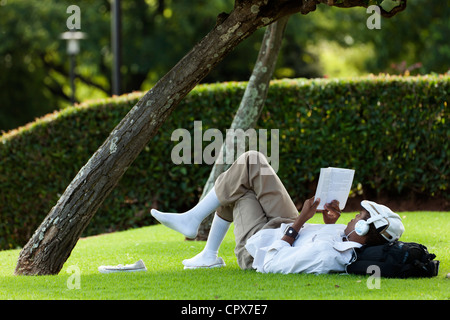 African man lying on grass, lire un livre tout en écoutant de la musique Banque D'Images