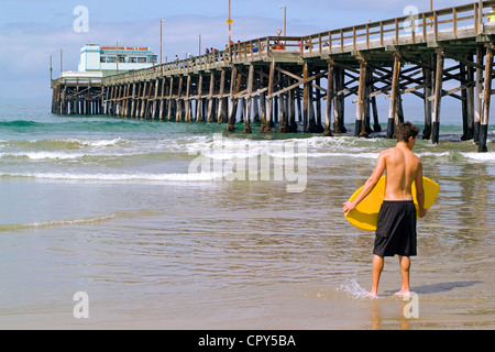 Un jeune skimboarder attend les conditions nautiques parfait comme les vagues de l'océan Pacifique en rouleau sous le Newport Pier à Newport Beach, Californie, USA. Banque D'Images