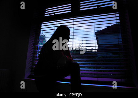 Jeune femme assise seule dans une pièce sombre à la recherche à travers une fenêtre aveugle. Banque D'Images