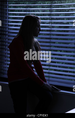 Jeune femme assise seule dans une pièce sombre à la recherche à travers une fenêtre aveugle. Propriété/modèle publié. Banque D'Images
