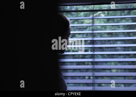 Jeune femme partiellement obscurci assis seul dans une pièce sombre à la recherche à travers une fenêtre aveugle. enfoncée sombre tristesse Banque D'Images