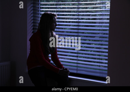 Jeune femme assise seule dans une pièce sombre à la recherche à travers une fenêtre aveugle. Banque D'Images