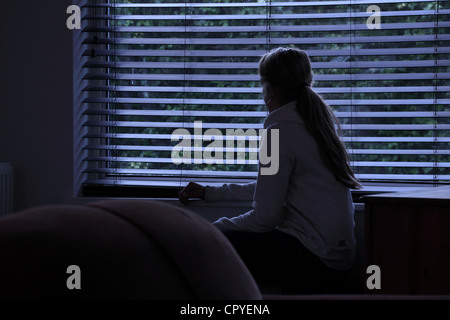 Jeune femme assise seule dans une pièce sombre à la recherche à travers une fenêtre aveugle. Banque D'Images