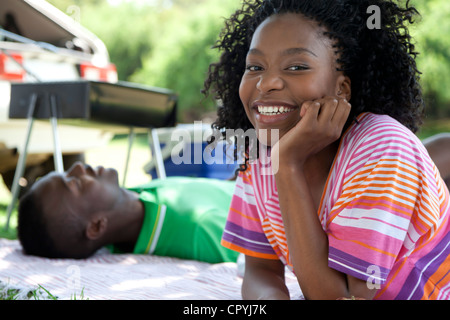 Gros plan du jeune femme noire appuyée sur sa main et smiling at camera, avec un jeune homme noir en arrière-plan de couchage Banque D'Images