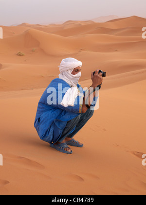 Homme touareg vêtu d'une robe bleue et d'un turban blanc de prendre une photo dans les dunes de sable du désert de l'Erg Chebbi Banque D'Images