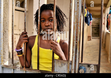 Fille avec un visage sérieux debout derrière une porte en treillis, Favela Morro da Formiga slum, district de Tijuca, Rio de Janeiro Banque D'Images