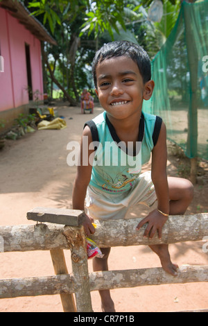 Smiling boy grimper sur une clôture avec leur mère assise derrière, Kanjippadom, près de Alappuzha (Alleppey), Kerala, Inde Banque D'Images