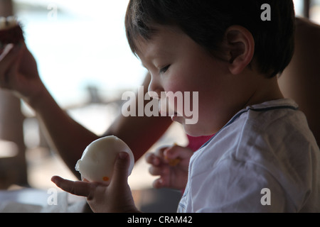 C'est une photo d'un petit garçon chinois qui est sur le point de mordre dans un morceau de pain. Il est très instable et semble faim Banque D'Images