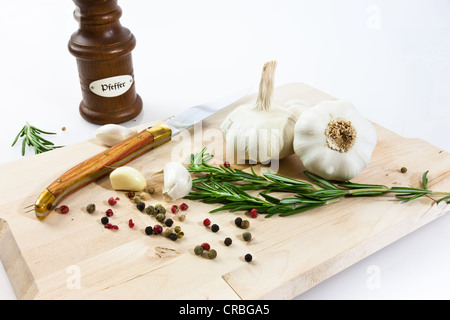 L'ail (Allium sativum) sur une planche en bois avec couteau, moulin à poivre, romarin et poivre noir et rouge Banque D'Images