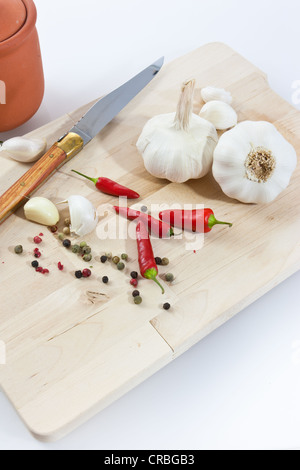 L'ail (Allium sativum) sur une planche en bois avec couteau, pot en argile, piment et poivre noir et rouge Banque D'Images