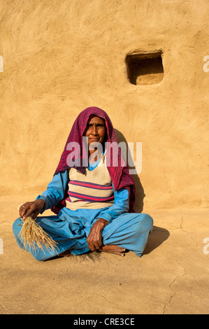 Femme âgée portant un foulard rouge tenant un balai dans sa main et assis devant le mur d'une maison, désert de Thar Banque D'Images