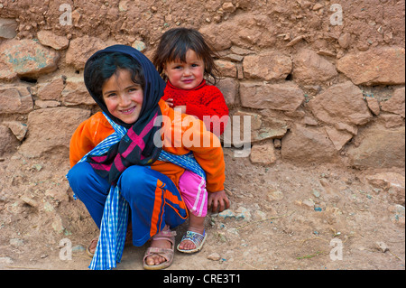 Jeune fille accroupie à un mur et sa jeune soeur dans un harnais sur son dos, ait Bouguemez, Haut Atlas Banque D'Images