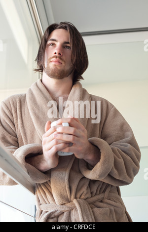 Young man holding mug en peignoir, près d'une fenêtre Banque D'Images