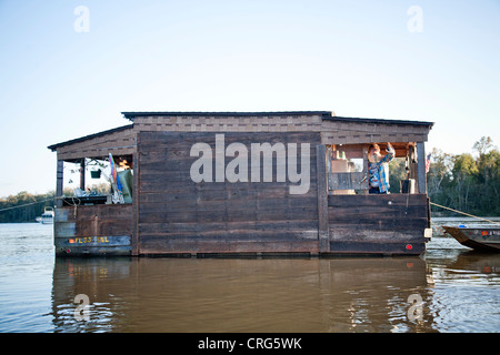 Un homme barbu se dresse sur le porche d'une petite péniche en bois avec un petit bateau de pêche attaché dans une rivière calme. Banque D'Images