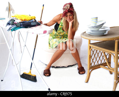 Jeune femme au foyer blonde de rêve avec un balai est assis entre planche à repasser avec pile de vêtements et une table avec des plats Banque D'Images