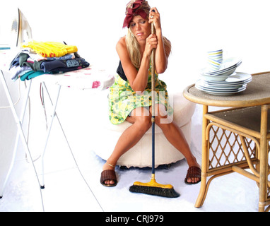 Fatigué et ennuyé blonde jeune femme au foyer avec un balai est assis entre planche à repasser avec pile de vêtements et une table avec des plats Banque D'Images