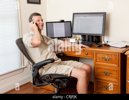L'homme travaillant à domicile bureau en short et d'un bureau avec deux moniteurs sur le téléphone Banque D'Images