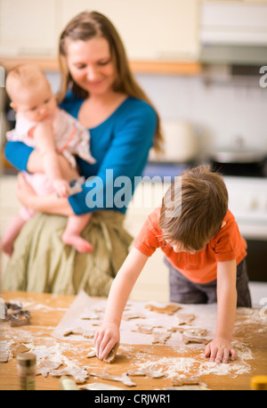 Jeune garçon dans la cuisine faire des cookies, smiling mother montres avec un bébé sur les bras Banque D'Images