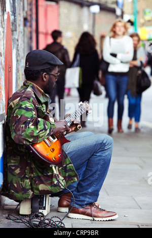 Portrait complet d'un busseur mâle assis sur la chaussée, jouant une guitare électrique, Brick Lane, Londres, Angleterre, Royaume-Uni. Banque D'Images