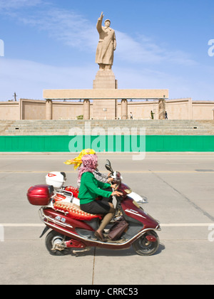 Avec le flou. 2 femmes sur les scooters chinois Ouïghours passer devant la statue de Mao Zedong à Kashgar. Banque D'Images
