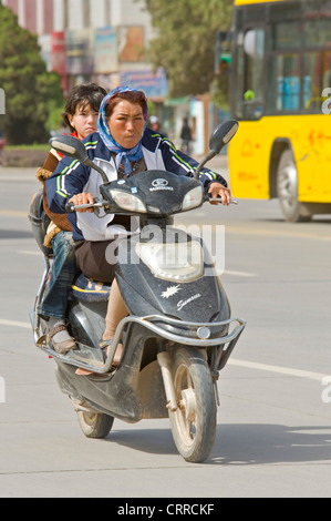 Une mère chinoise et sa fille voyagent le long d'une route sur leur scooter dans Kashgar. Banque D'Images