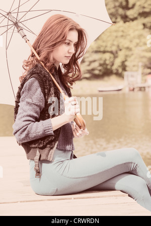 Jeune femme avec parapluie blanc sitting by a lake looking pensive Banque D'Images