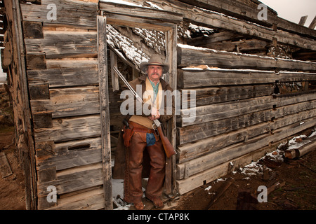 Old cowboy appuyé contre un cadre de porte d'une vieille cabane dans la tenue d'hiver son fusil Banque D'Images