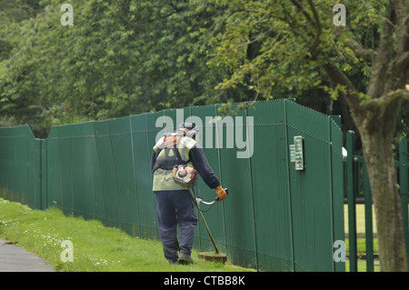 Travailleur du conseil strimming herbe dans le parc local par une clôture Banque D'Images