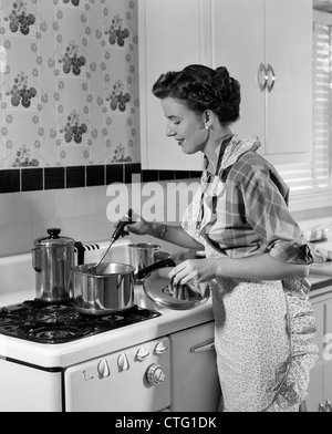 Années 1950, maîtresse de l'ALIMENTATION DANS UNE CASSEROLE EN REMUANT TABLIER SUR Cuisinière Banque D'Images