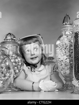 1950 SMILING GIRL avec bonnet et gants blancs pris en sandwich entre les grands pots de bonbons SUR LE COMPTOIR Banque D'Images