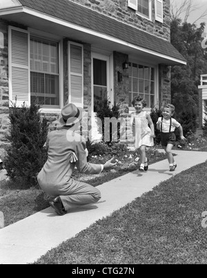 1950 HOMME PÈRE BRAS agenouillé devant la maison de banlieue pour saluer BONNE COURSE BIENVENUE Accueil fils et fille Banque D'Images