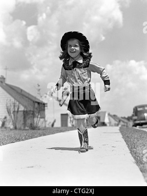 1950 FILLE EN COSTUME COWGIRL FONCTIONNANT SUR TROTTOIR VERS LA CAMÉRA Banque D'Images