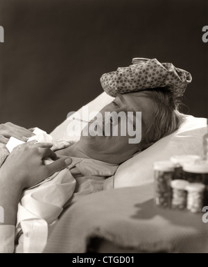 Années 1960 Années 1970 DANS L'HOMME MALADE AU LIT AVEC LE THERMOMÈTRE DANS LA BOUCHE DE GLACE SUR LA TÊTE DE BOUTEILLES DE MÉDICAMENT À PROXIMITÉ Banque D'Images