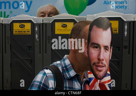 Avec un organisme de bienfaisance billboard derrière, un spectateur porte un masque avec le visage de l'équipe Go de cyclisme sur route de héros Mark Cavendish sur le premier jour de compétition de l'Olympique de Londres 2012 250km course sur route des hommes. À partir du centre de Londres et le passage du capital des sites célèbres avant de partir dans l'Angleterre rurale à l'épuisant fort Hill dans le comté de Surrey. Sud-ouest locaux Londoniens étaient alignés sur la route dans l'espoir de la Cavendish favori pour gagner la médaille de l'équipe Go premier mais ont finalement été déçu quand le Kazakhstan's Alexandre Vinokourov a finalement remporté l'or. Banque D'Images