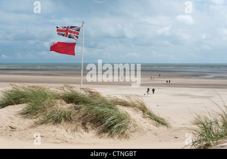 Union jack flag et red flag flying des dunes sur la plage par un jour de vent à marée basse Banque D'Images