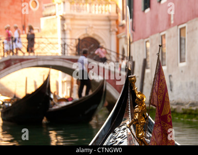 Vue depuis le bord d'un canal typique le long de gondole avec proue de bateau, gondolier, gondoles et les gens sur le pont Venise Vénétie Italie Banque D'Images