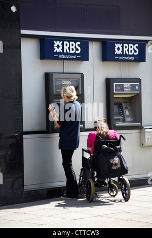 Jeune femme à l'aide de RBS International cash machine tandis qu'un autre attend aux côtés de fauteuil roulant à St Helier, Jersey en Juillet Banque D'Images