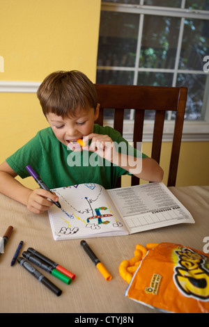 Un garçon mexicain-américain de sept ans goûte à la malbouffe, Cheetos, tout en dessinant à la maison avec des marqueurs. Banque D'Images