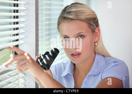 Woman peering through blinds avec des jumelles Banque D'Images