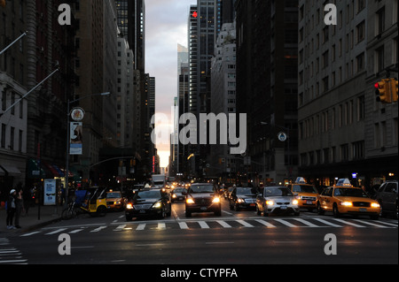 Coucher du soleil 'urban alley' de la 6e Avenue à plusieurs voies de circulation phares, voitures, taxis, l'article 59e Rue Ouest intersection, New York Banque D'Images